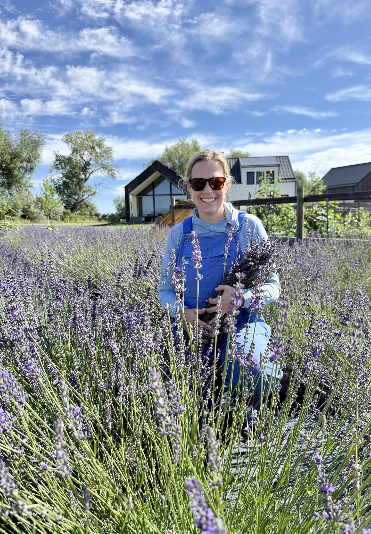 Laura smiling in the lavender rows, holding a freshly cut bundle, with the farmhouse behind her